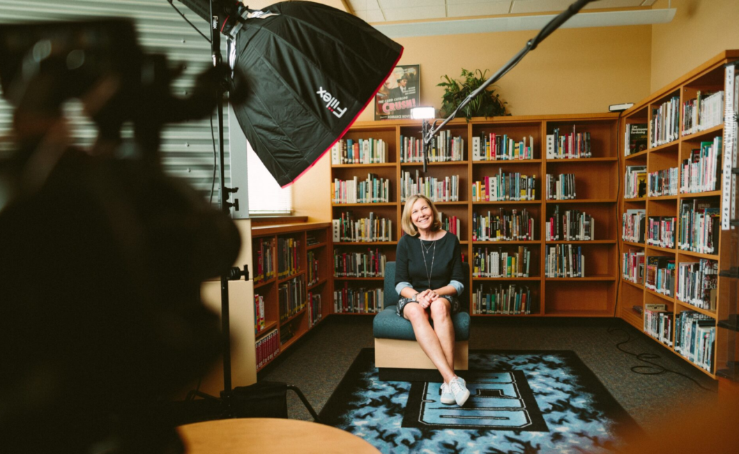 Woman sitting in front of bookshelves and smiling while being recorded by a film crew