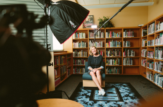 Woman sitting in front of bookshelves and smiling while being recorded by a film crew