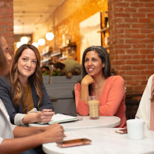 A group of four professional women sitting around two white tables in a coffee shop